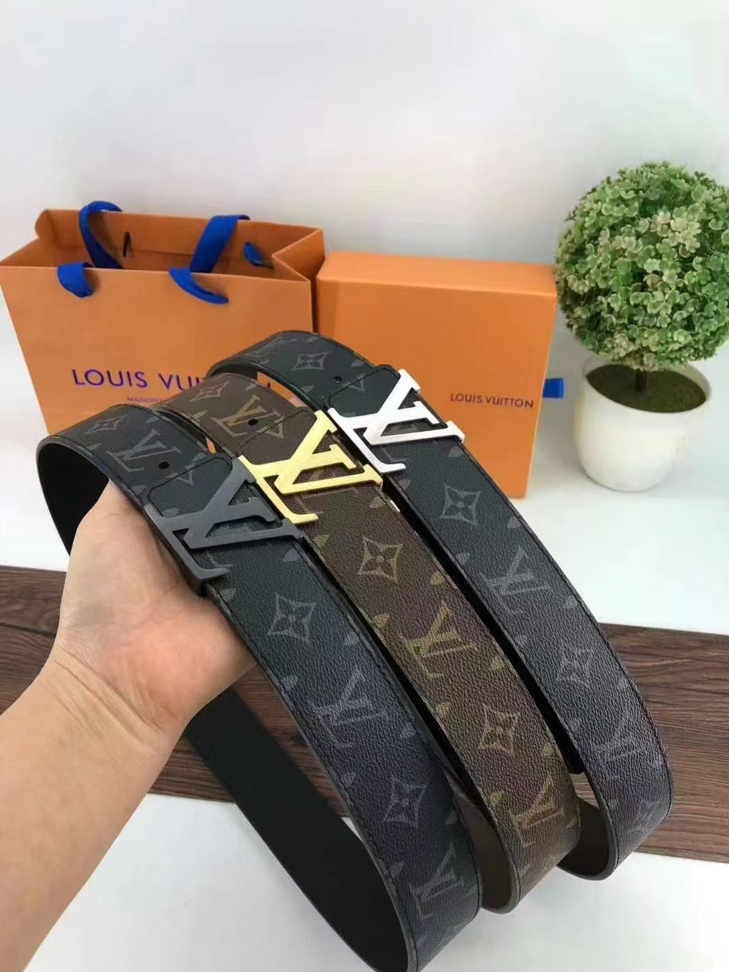 Louis Vuitton belts with monogram pattern held by a hand, with brand packaging and a plant in the background.